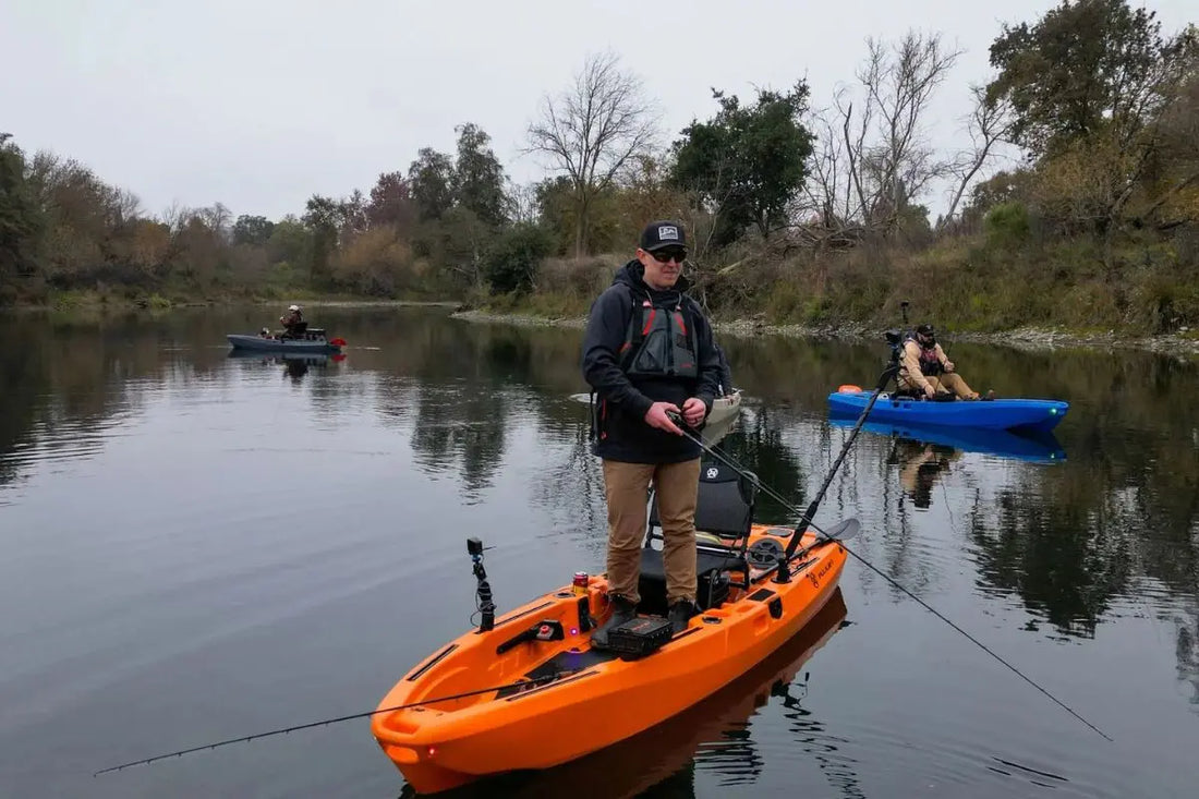 Angler standing up to fish on an orange FluxJet electric jet kayak in a river, demonstrating hull stability with blue and grey models in the background.