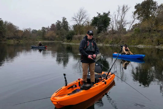 Angler standing up to fish on an orange FluxJet electric jet kayak in a river, demonstrating hull stability with blue and grey models in the background.