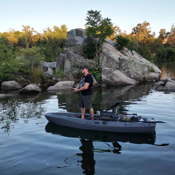 Angler standing on a gray FluxJet electric kayak beside rocks and trees on a calm lake