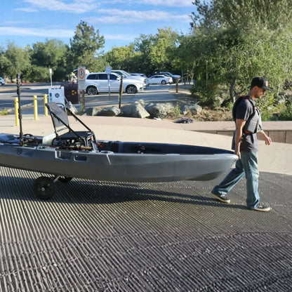 Man pushing a large kayak on wheels along a paved path with trees and cars in the background.