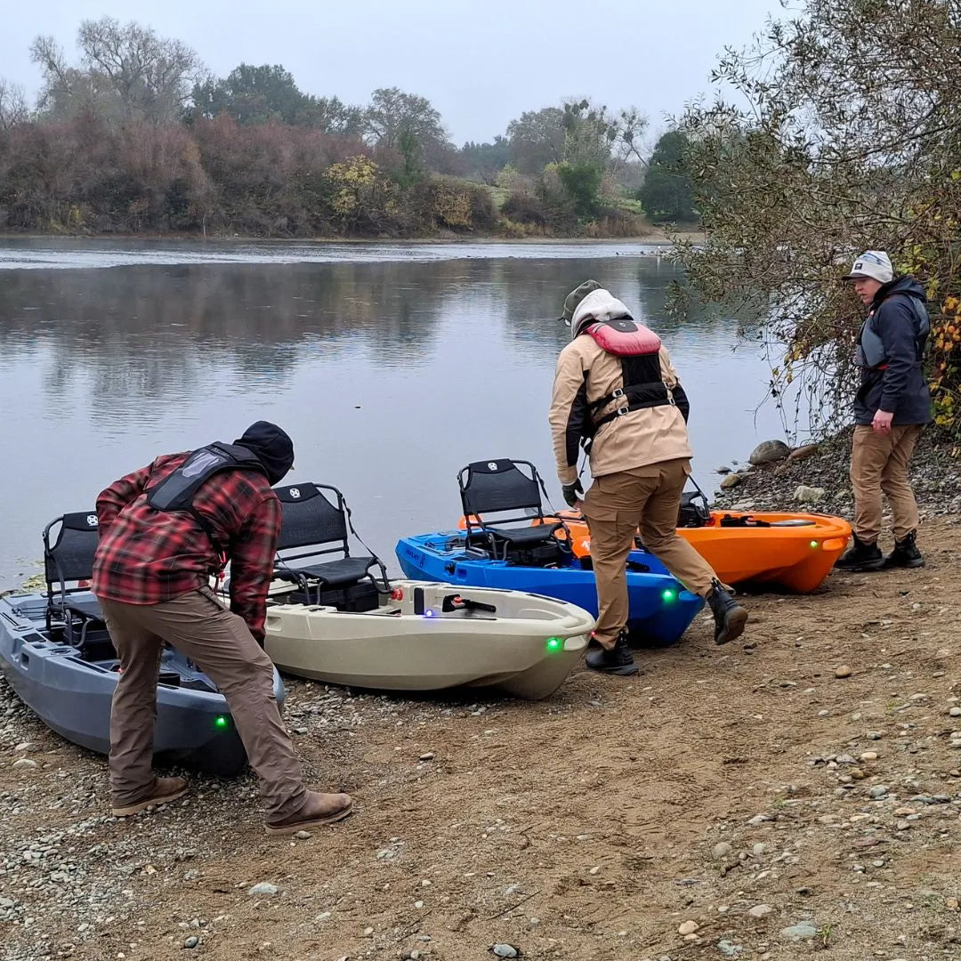  People preparing and positioning multiple FluxJet kayaks on a rocky riverbank before launching into calm water.